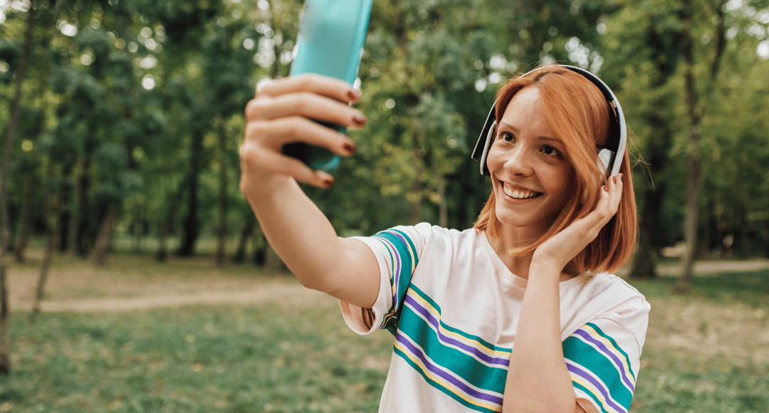 Rødhåret smilende pige med headset på, der holder sin telefon op i luften men hun facetimer fra sin nye mobilpakke hos velkommen 