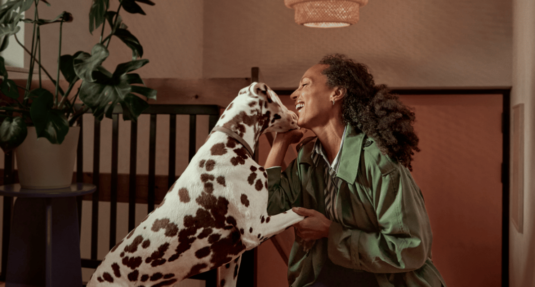 Dame med mørkt krøllet hår og grøn jakke, der bliver budt velkommen hjem til god energi af glad hund