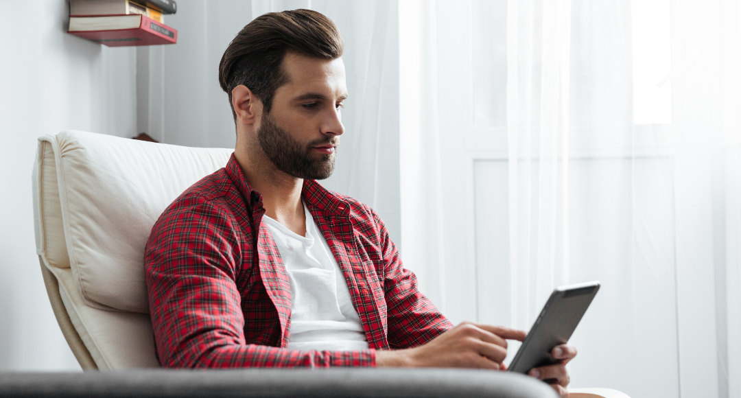 Mand med mørkt hår og skæg og rød skjorte, sidder i lænestil med mørk tablet i hånden og har lige bestilt mobilt bredbånd hos Velkommen