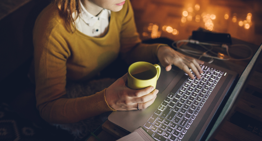 Kvinde med skjorte og gul trøje og en gul kop i hånden tjekker sin elregning fra velkommen på sin computer .png