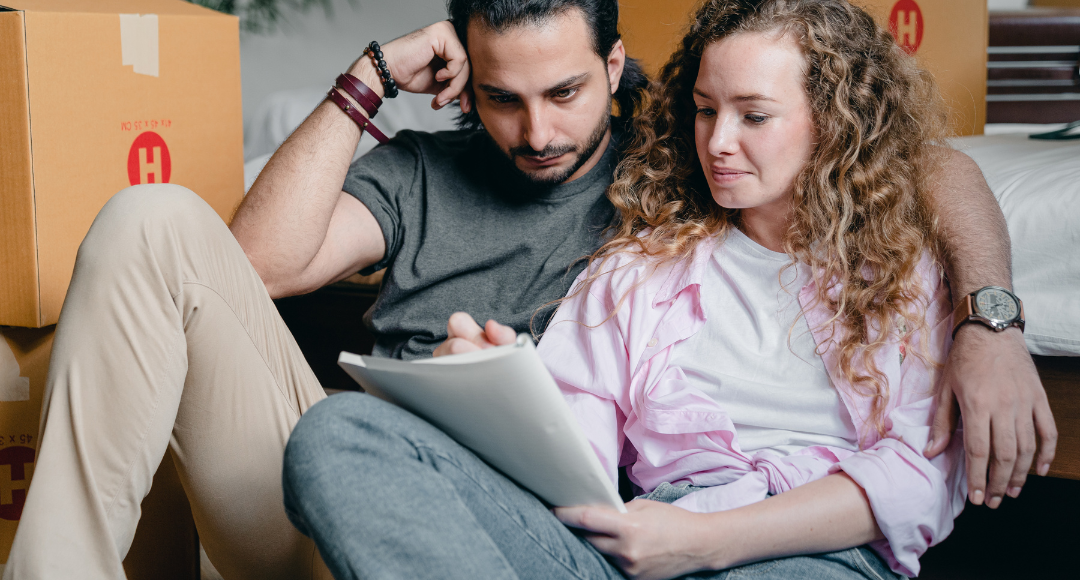 Mand og kvinde sidder på gulvet blandt flyttekasser. Manden har armen om kvinden, der sidder og noterer på en papirblok, hvad de skal have styr på under flytningen. 