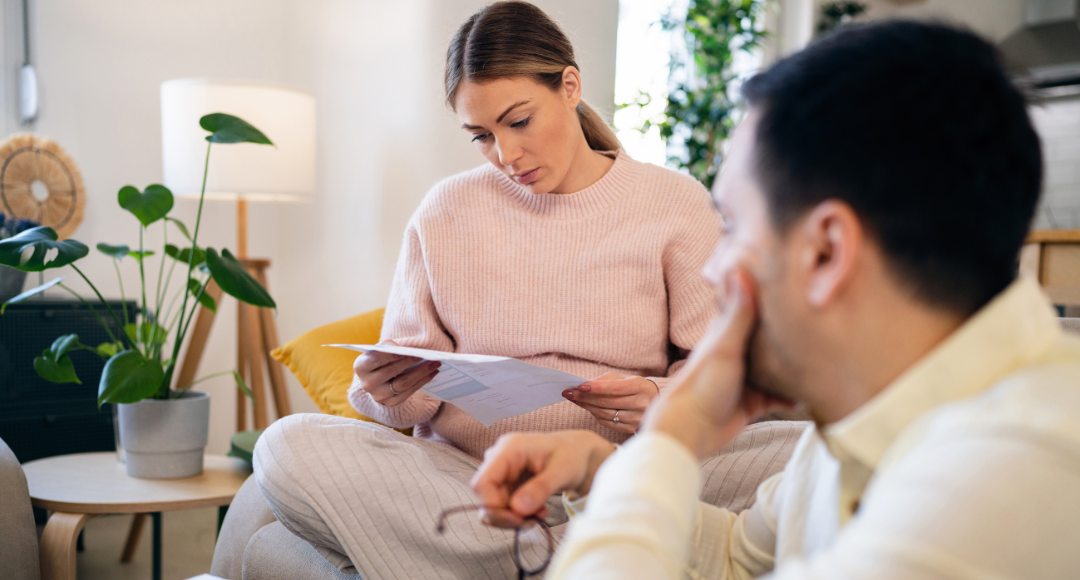 kvinde i lyserød sidder og kigger på sin regning og mand sidder i hvid og holder hånden til ansigtet med briller i hånden 